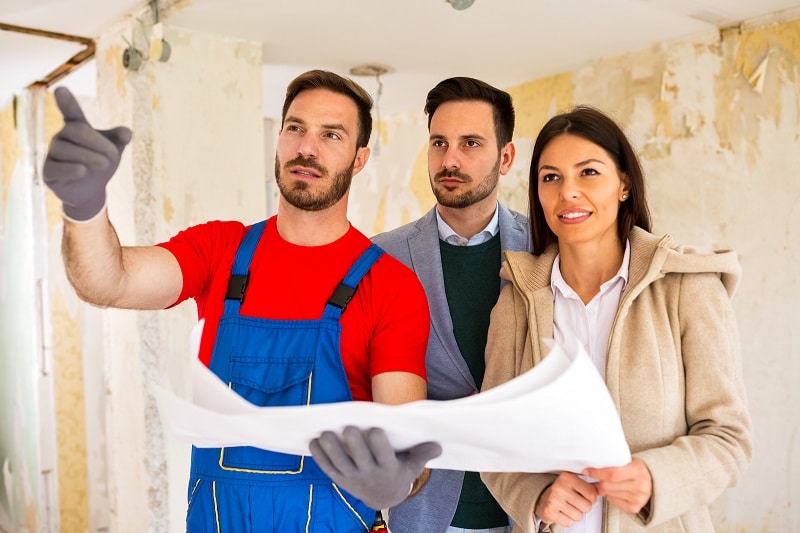A contractor holds on to housing blueprints as he points something out in a house undergoing a major renovation. A couple looks on.