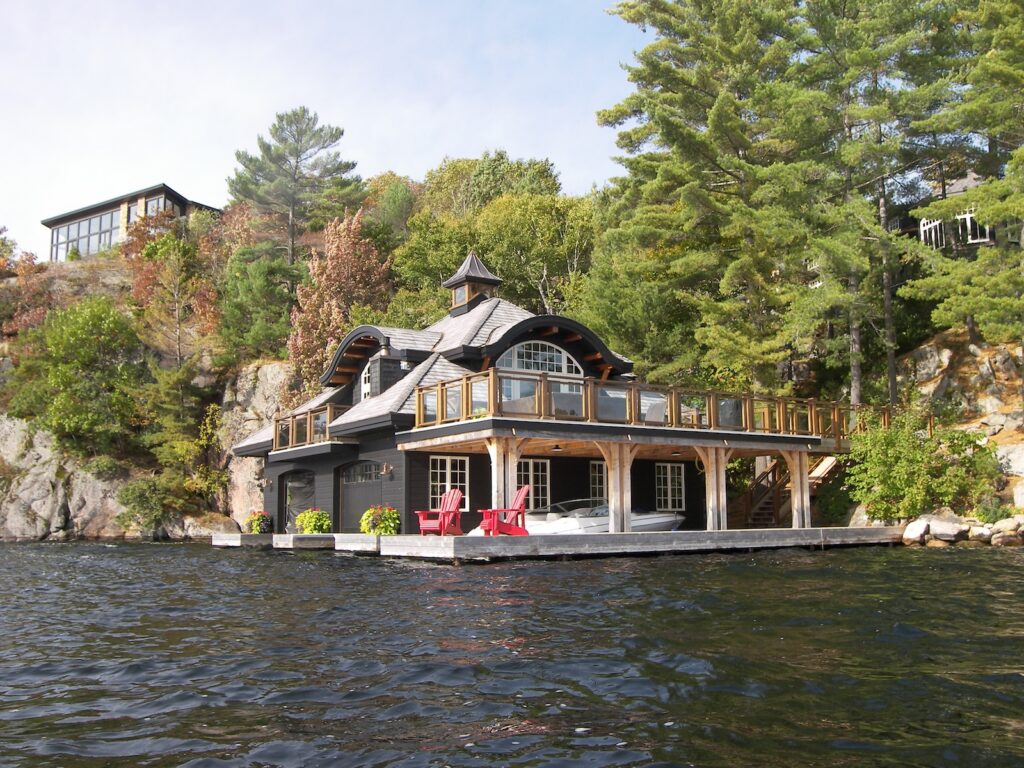 A luxury black boathouse features a peaked roof and a spacious second-story deck above the dock and boat slips.