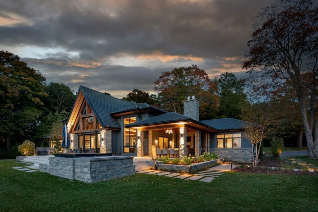 Custom home renovation with stone exterior, modern lighting, and covered patio surrounded by trees at sunset in Cottage Country.