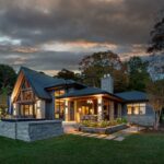 Custom home renovation with stone exterior, modern lighting, and covered patio surrounded by trees at sunset in Cottage Country.