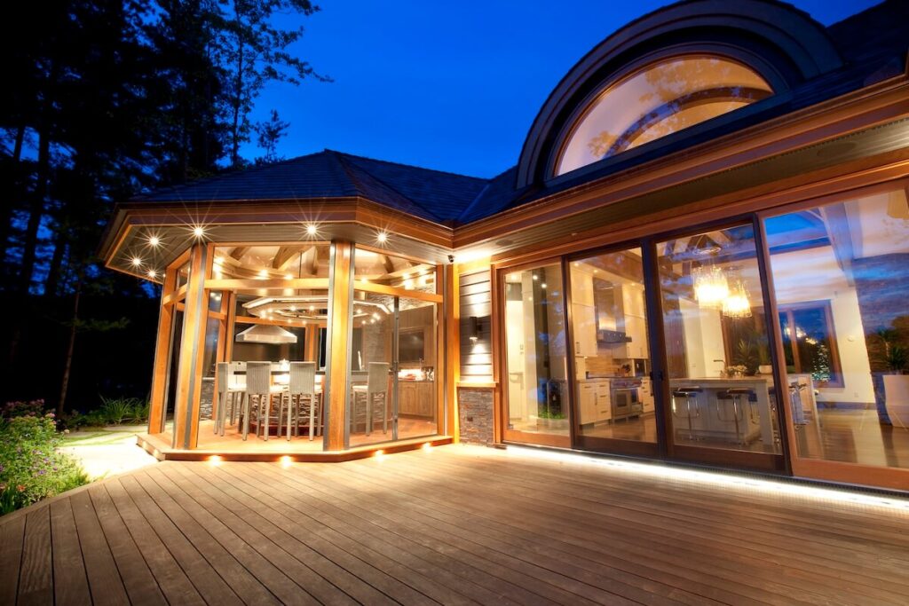 Luxury custom home at dusk with glowing exterior lights, expansive deck, and glass-enclosed kitchen and dining pavilion nestled in nature.