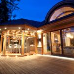 Luxury custom home at dusk with glowing exterior lights, expansive deck, and glass-enclosed kitchen and dining pavilion nestled in nature.