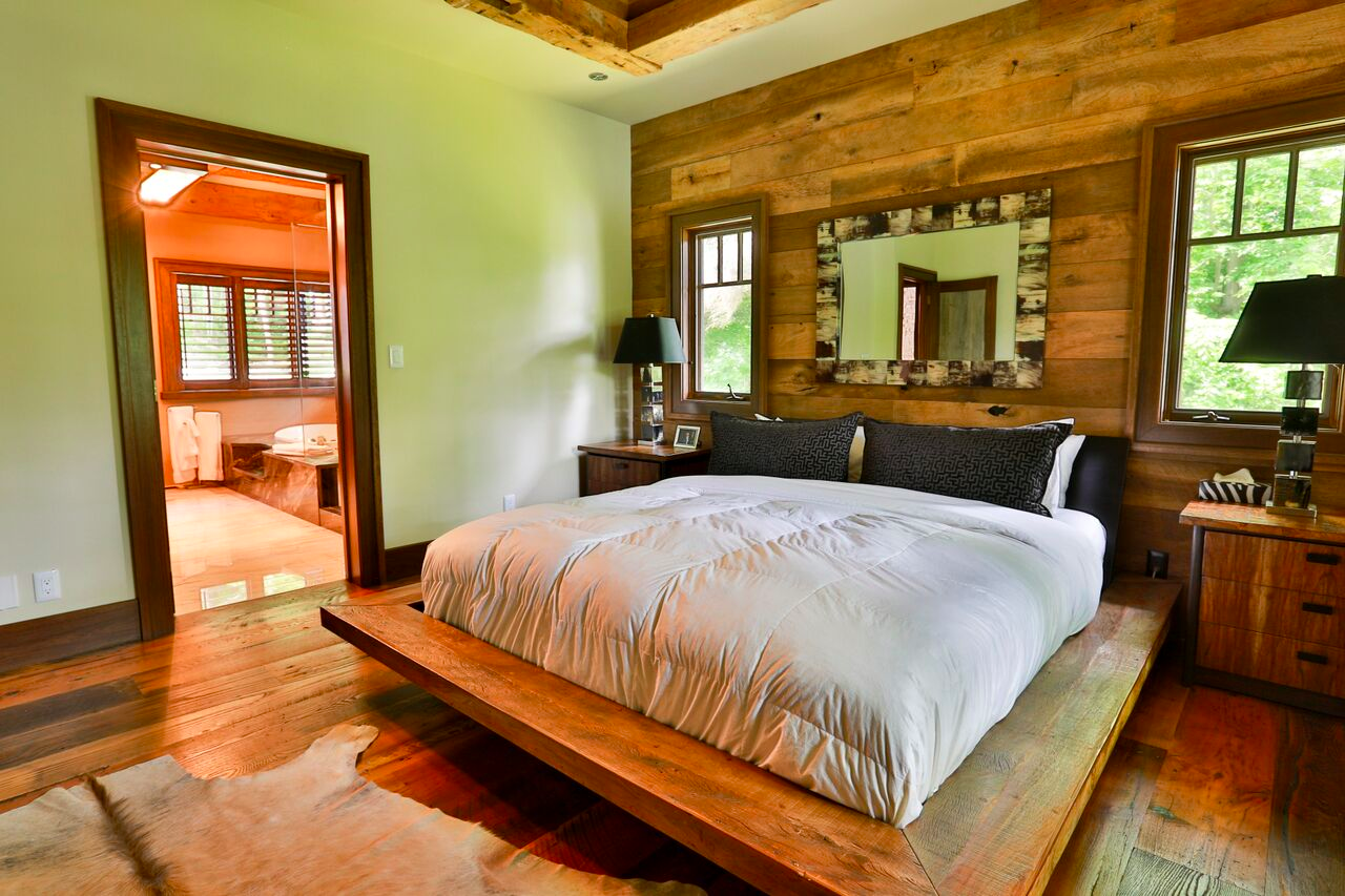 A cozy, rustic bedroom with reclaimed wood on the floor, one of the walls, and in the bedframe. The view includes an open door towards the master bathroom.