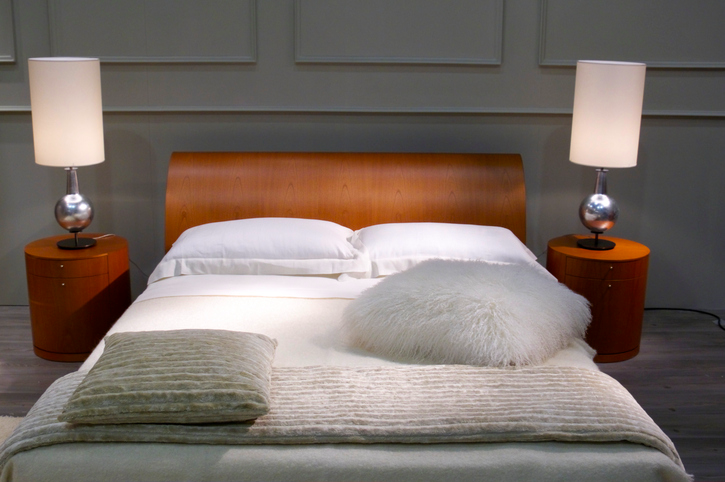 A view towards a bed with a stained wood headboard. There is a circuler stained wood table on each side of the bed, with sleek, cylindrical metal lamps on each table.