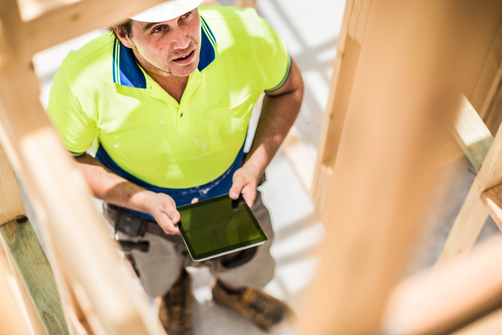 A contractor holds on to a tablet as he looks up towards the building frame of a new home.