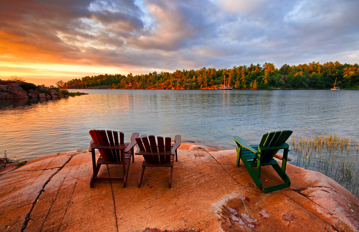 A few Muskoka chairs sit on a rocky outcrop, overlooking an Ontario lake during sunset.