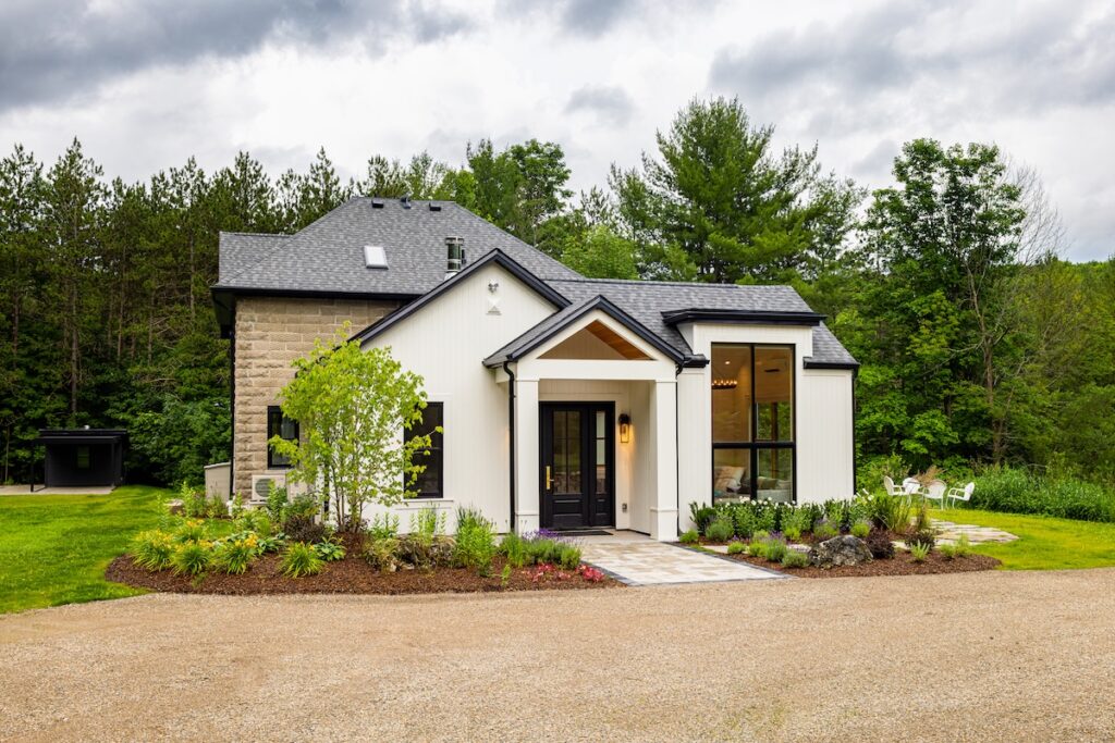 A modern white cottage with black trim and landscaped gardens set against a forest backdrop in Muskoka.