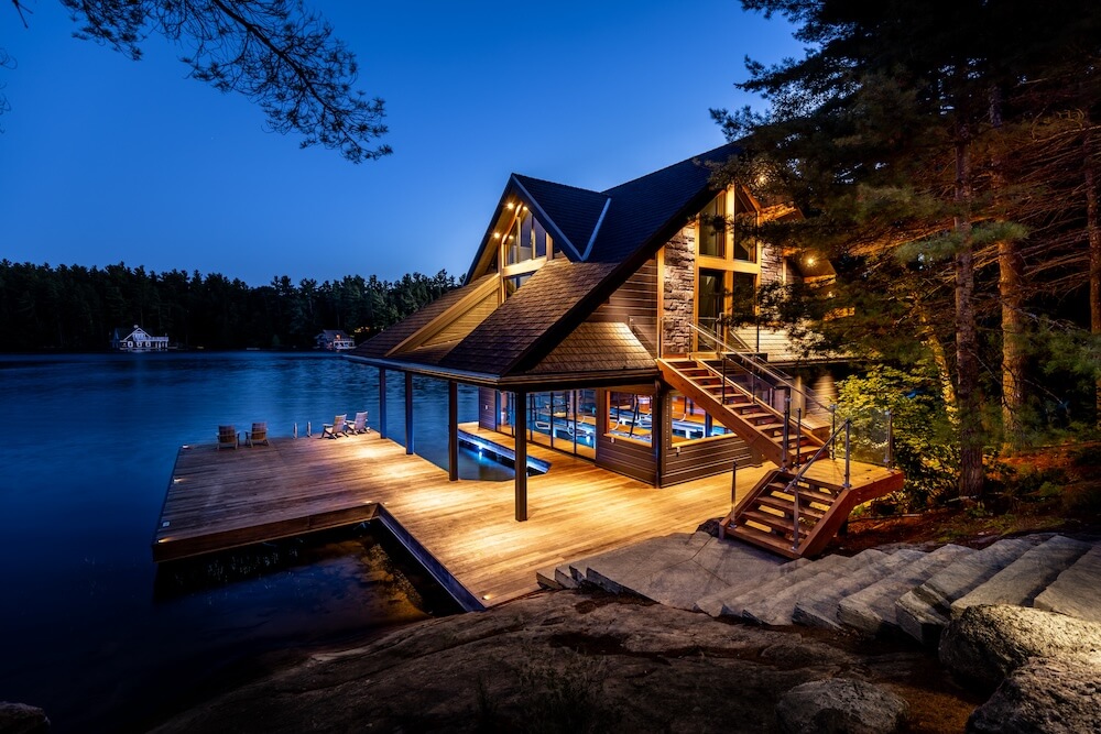 A luxurious custom boathouse at dusk, featuring a two-level design with large windows, natural stone accents, and wood siding. The structure extends over the water with integrated boat slips below and a spacious wraparound dock above, lined with Adirondack chairs. Surrounded by pine trees and lit with warm, ambient lighting, the boathouse sits on a quiet lake in Ontario’s Cottage Country.