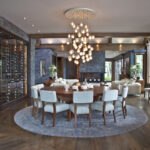 An elegant dining room featuring a large wooden table surrounded by light blue upholstered chairs. Above the table hangs an impressive chandelier with multiple clear orbs casting a warm glow. The room has rich hardwood flooring and stone columns with a slate blue tone that match a wall wine rack enclosure. In the background, there's a view into an adjacent room with matching decor, continuing the luxurious yet welcoming ambiance.