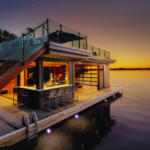 Luxury two-story Muskoka boathouse with rooftop deck, glass railings, and warm interior lighting at sunset.