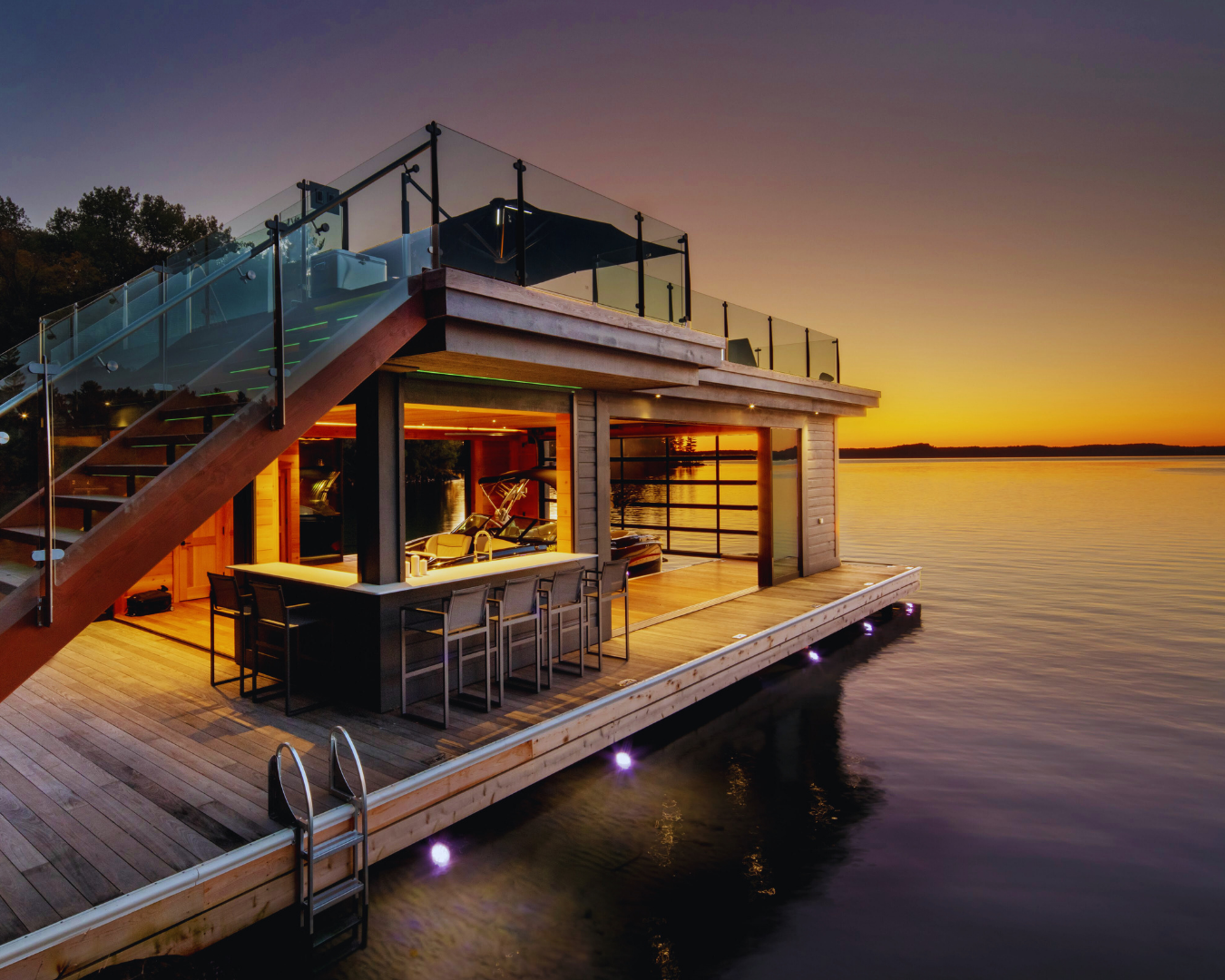 Luxury two-story Muskoka boathouse with rooftop deck, glass railings, and warm interior lighting at sunset.
